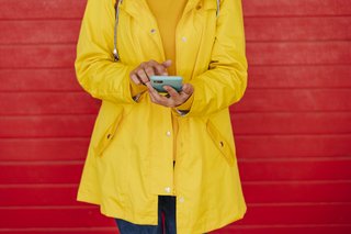 Eine Frau in einem gelben Regelmantel steht vor einer roten Steinmauer, tippt etwas in ihr Smartphone.