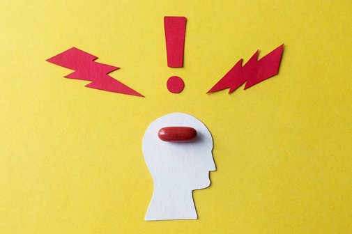 Symbol f&uuml;r Migr&auml;ne und Kopfschmerzen: Ein aus Pappe ausgeschnittener stilisierter Kopf auf gelben Hintergrund, darin liegt eine rote Pille. Um den Kopf herum symbolisieren zwei rote Blitze und ein rotes Ausrufezeichen Schmerzen.