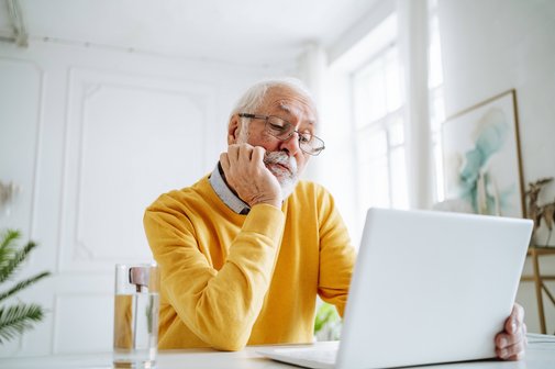 Ein Senior mit Brille im gelben Pullover sitzt an einem Tisch und schaut auf ein Notebook. Das Kinn hat er in die Hand gestützt.