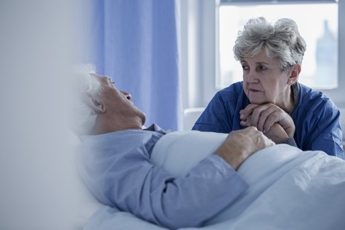 Ein alter Mann liegt in einem Bett in einem Krankenhaus oder Pflegeheim, eine Frau sitzt am Bett und hält seine Hand.
