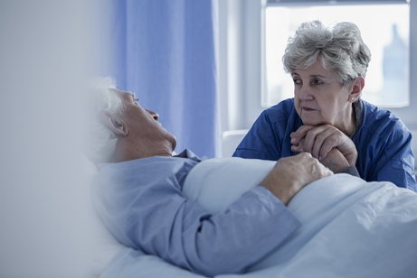 Ein alter Mann liegt in einem Bett in einem Krankenhaus oder Pflegeheim, eine Frau sitzt am Bett und h&auml;lt seine Hand.