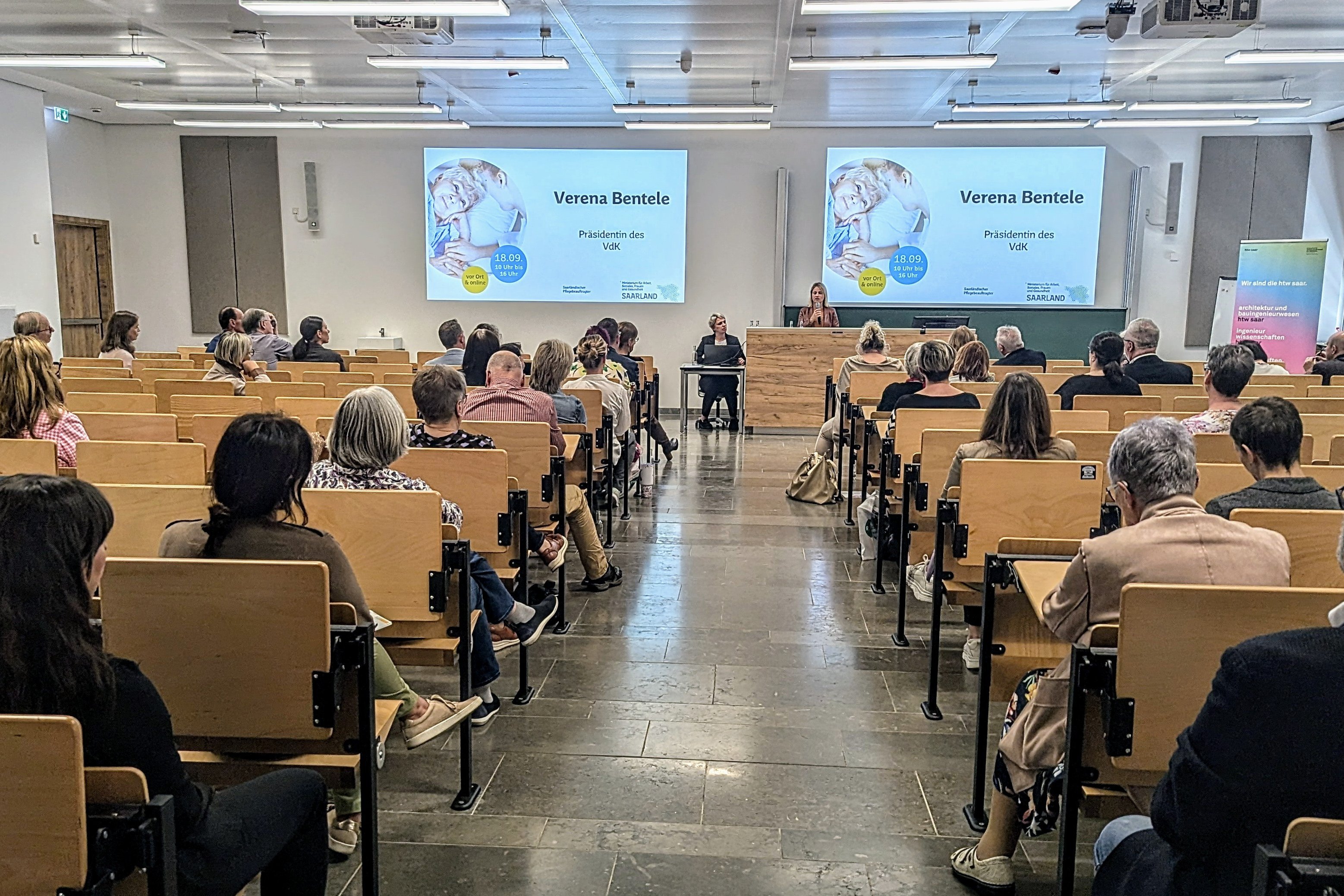 Zuhörende sitzen in einem Hörsaal der HTW, hinten spricht VdK-Präsidentin Verena Bentele.