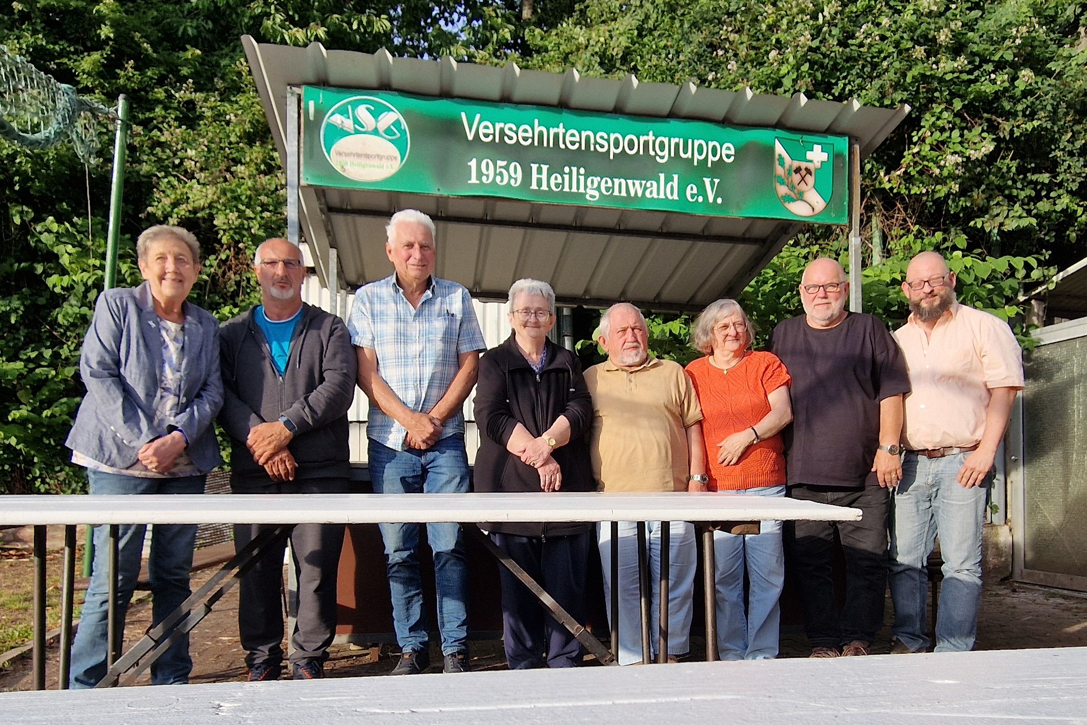 Auf dem Photo ist der im April 2026 gew&auml;hlte Vorstand des VdK-Ortsverbandes Heiligenwald abgebildet. Diese drei Frauen und f&uuml;nf M&auml;nner stehen von der Sonne angestrahlt vor einem Grillstand der Versehrtensportgruppe 1959 Heiligenwald e.V.. Im Hintergrund sind gr&uuml;ne Laubb&auml;ume zu sehen.